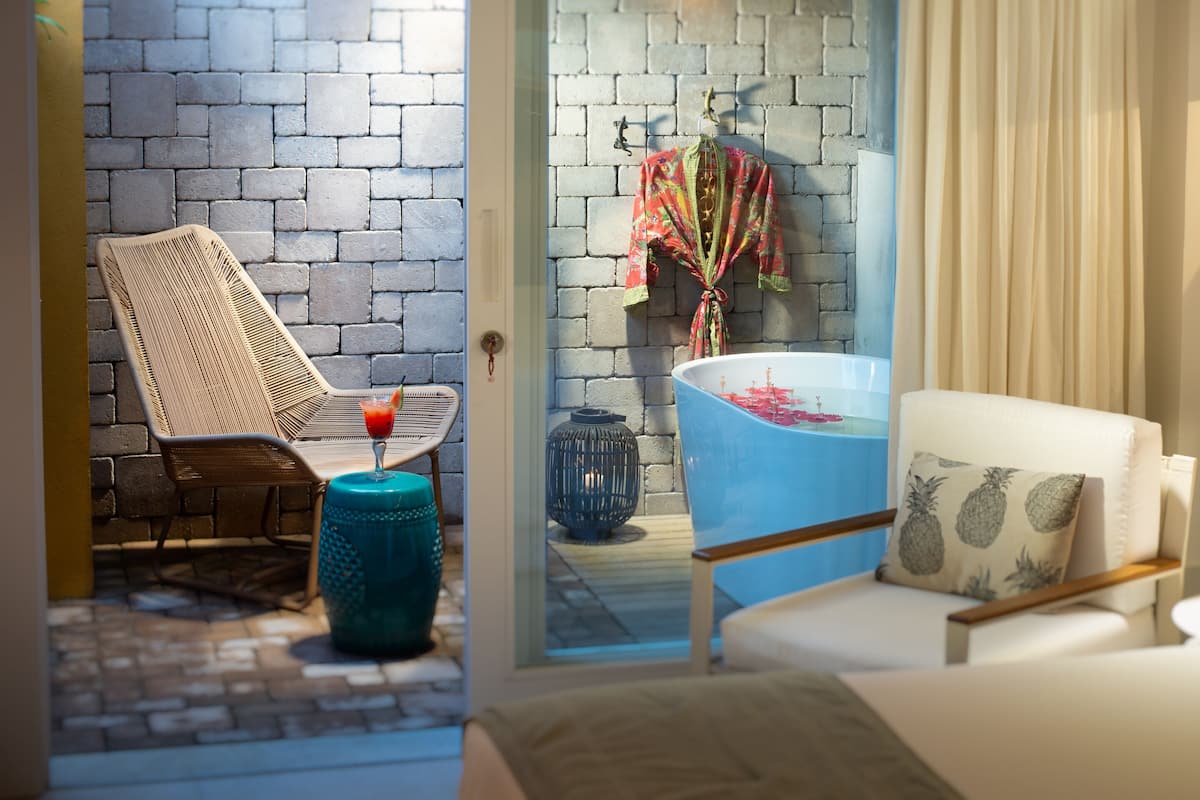 A hotel room with a brick wall, a wicker chair, a blue bathtub, a white sofa, and a floor lamp.