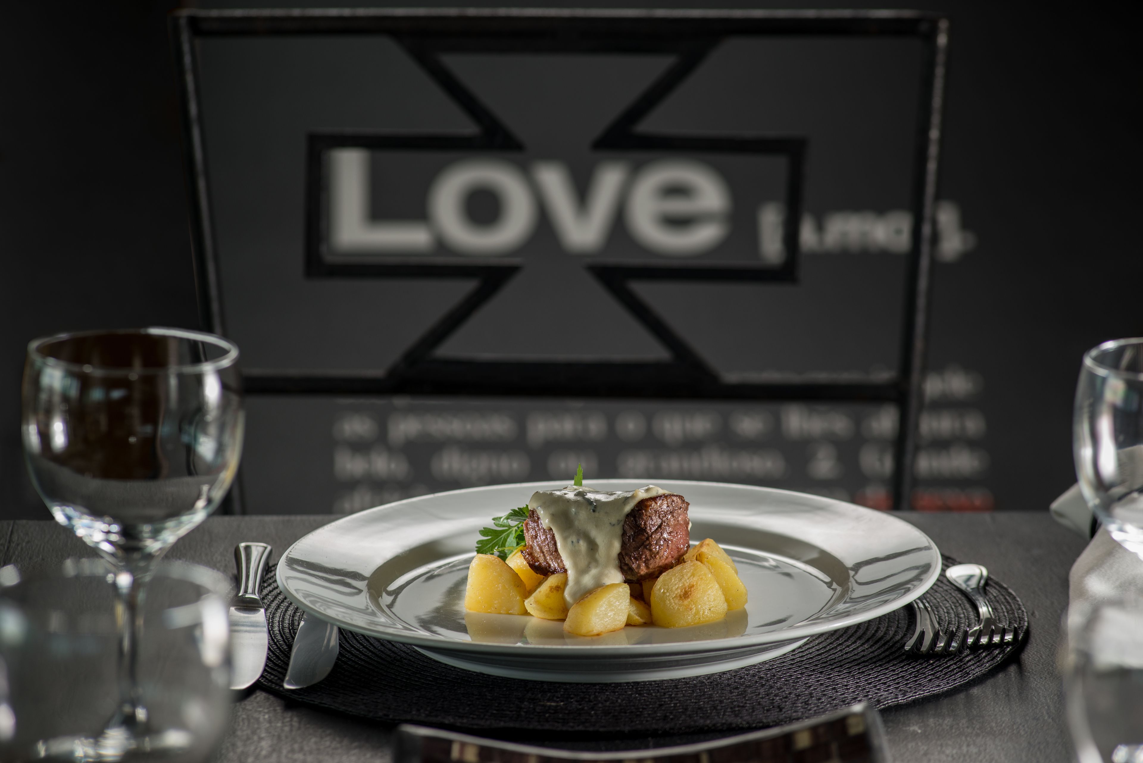 Une assiette de nourriture comprenant un steak et des pommes de terre, un verre de vin et un panneau noir sur lequel on peut lire le mot « Amour » en arrière-plan.