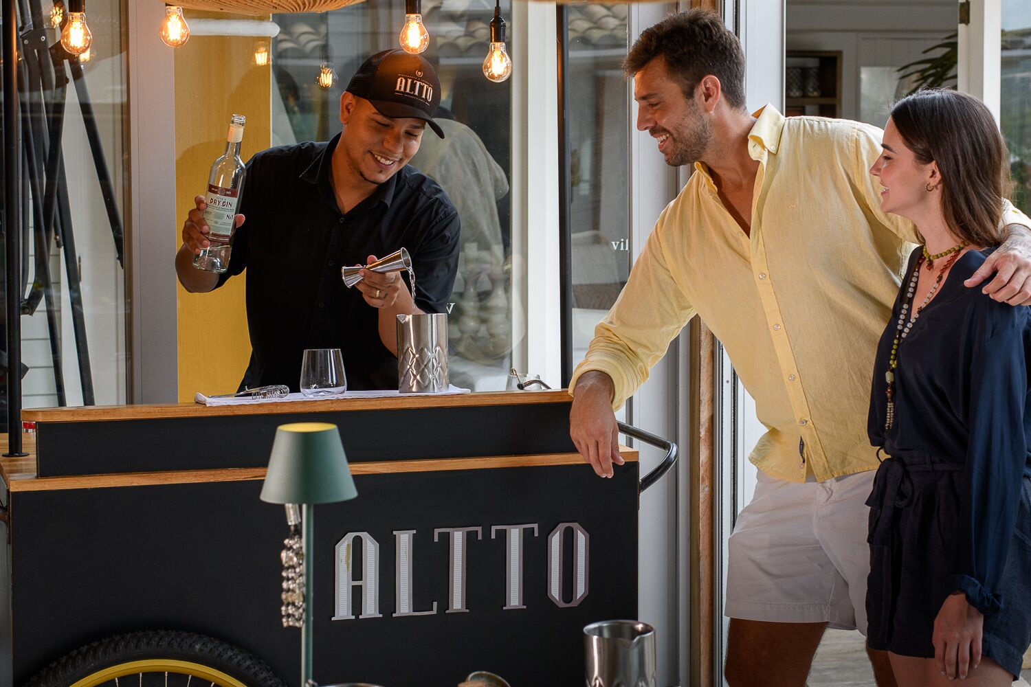 A bar area with a counter labeled "ALTTO," a person pouring a drink, and a woman standing nearby.