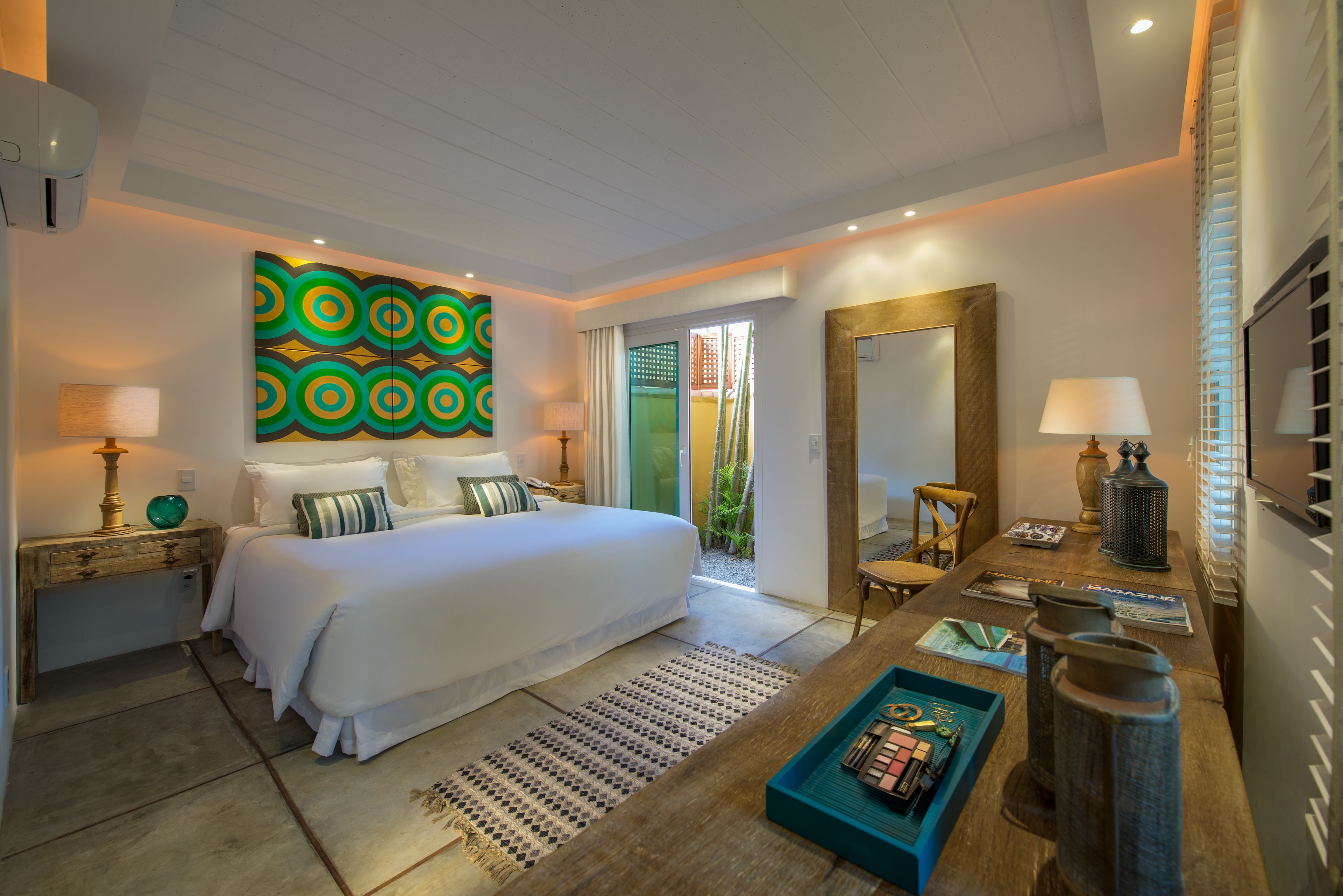 A modern hotel room with a large bed, a wooden nightstand, a desk with a lamp, a mirror, and a colorful abstract painting on the wall.