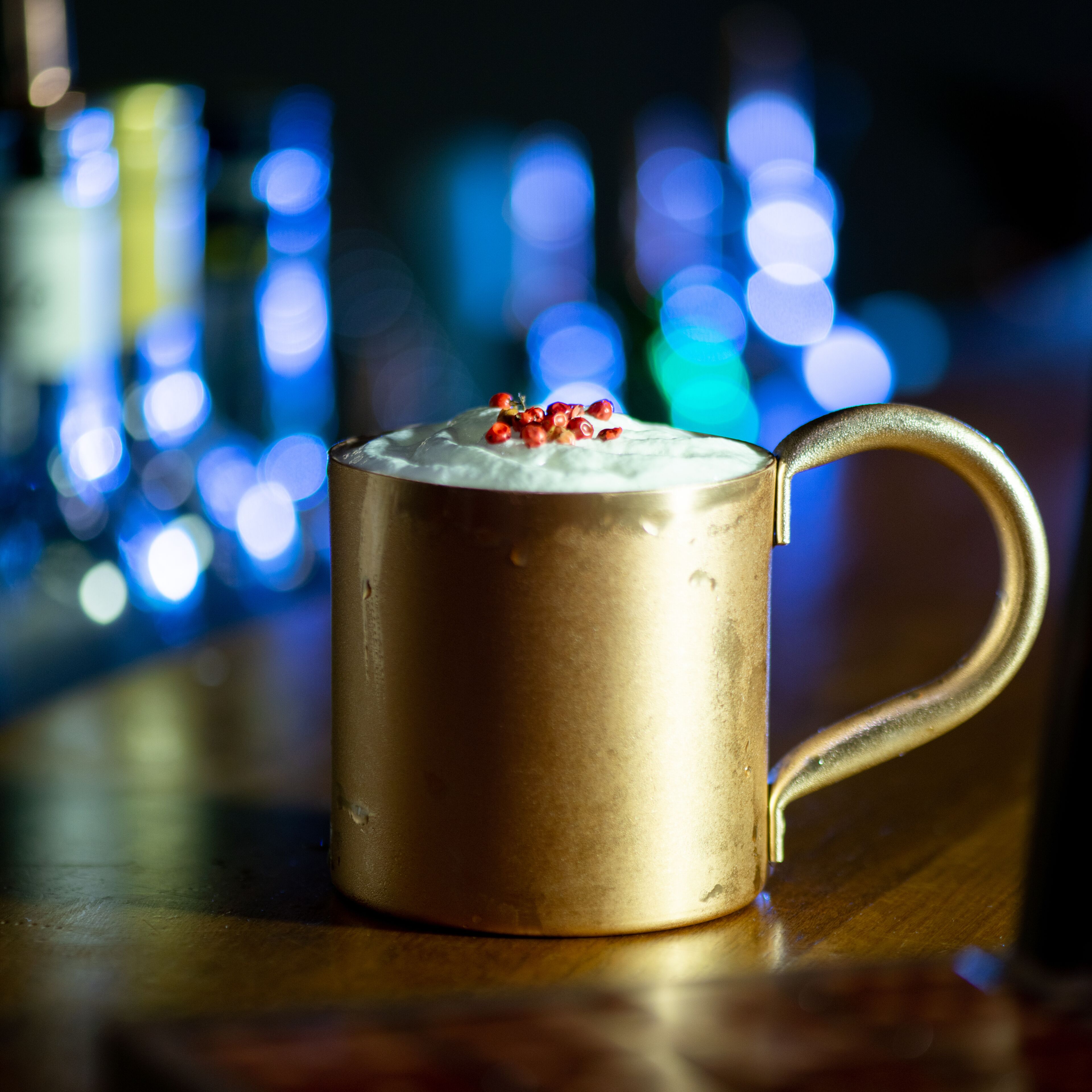 Una taza dorada con asa, llena de una bebida cremosa y adornada con bayas, sobre una superficie de madera con luces desenfocadas al fondo.