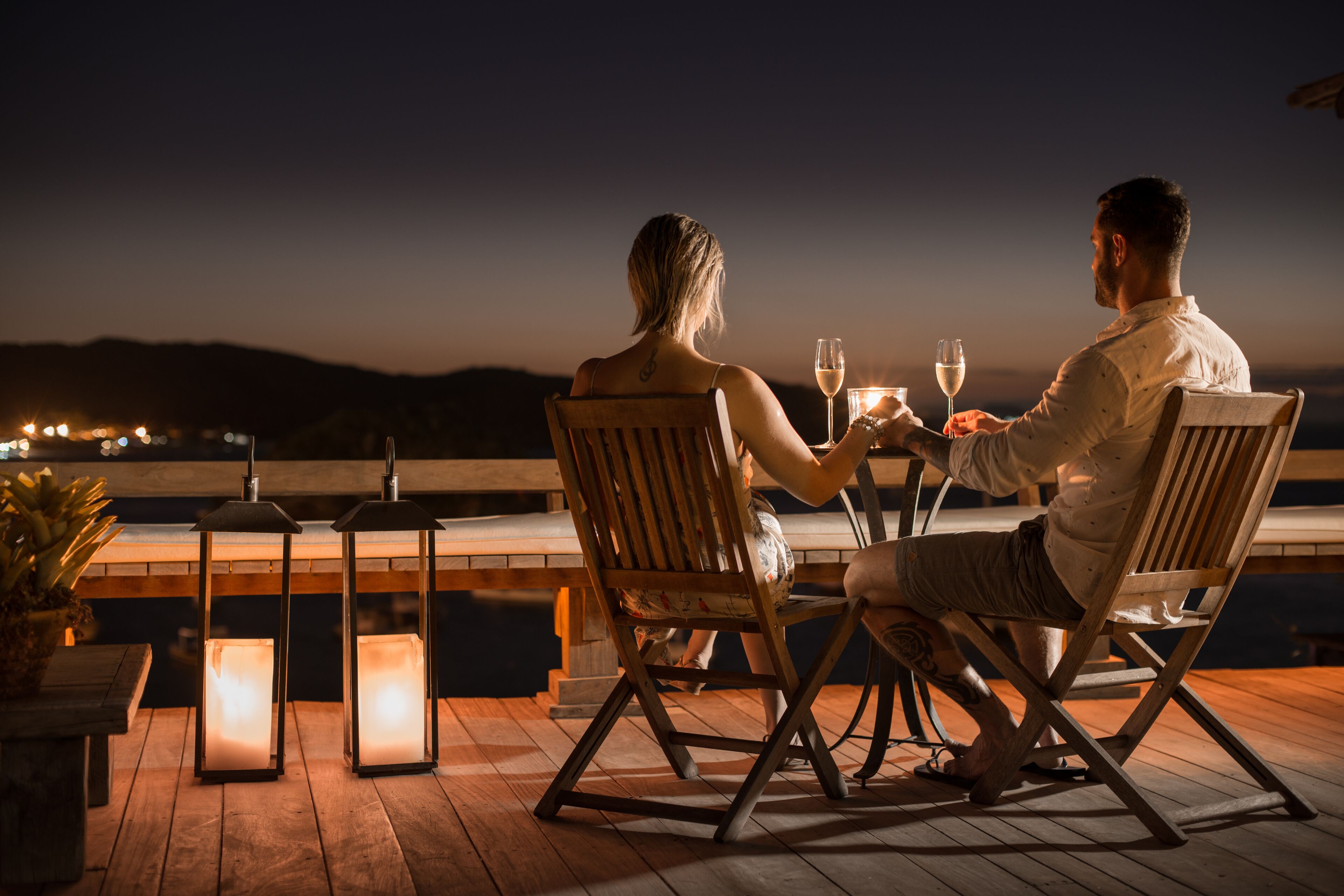 A couple dining at a wooden table with lanterns on a deck overlooking a body of water at dusk.