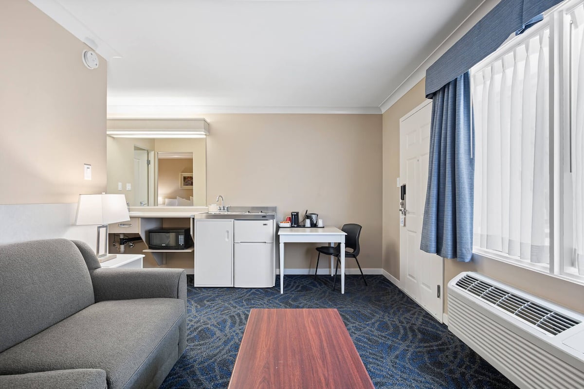 Ett hotellrum med en soffa, ett litet skrivbord med en stol, ett litet kök, en spegel och ett stort fönster med gardiner.