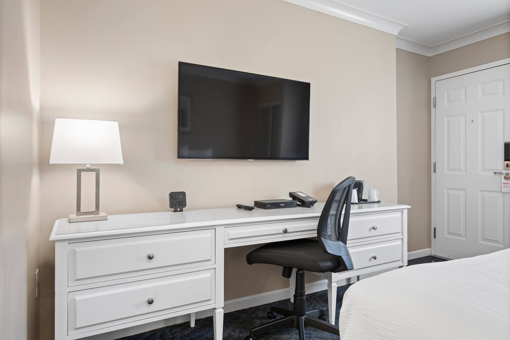 Ett hotellrum med ett vitt skrivbord, en svart kontorsstol, en platt-TV som är monterad på väggen, ett sängbord med en lampa och en säng med ett vitt överkast.