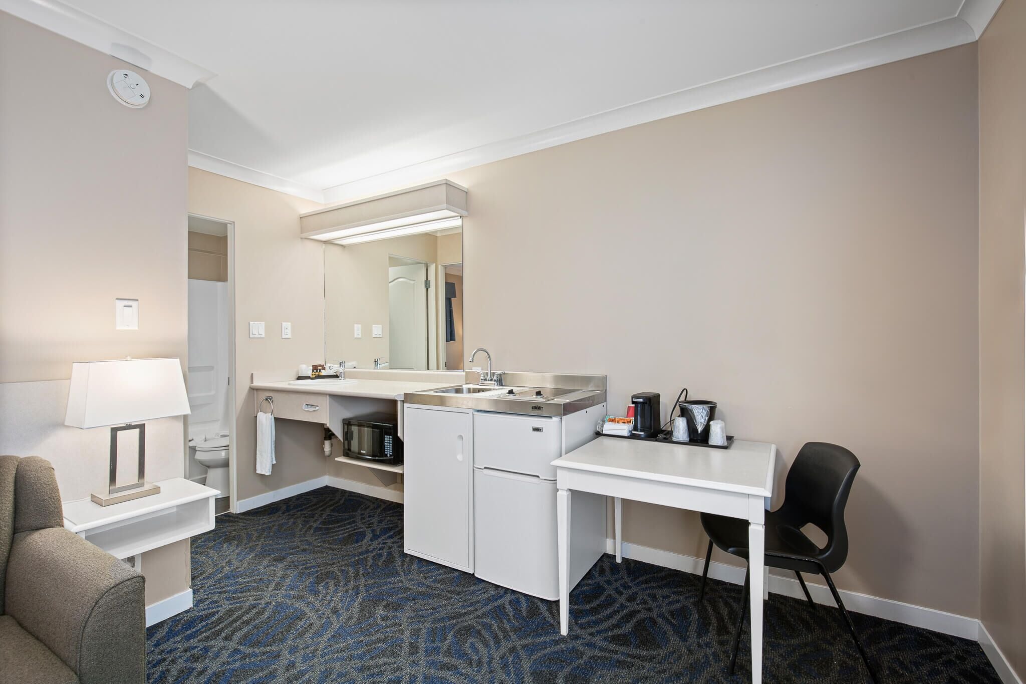 Habitación de hotel pequeña con kitchenette, una cama individual, escritorio con silla y baño con espejo y lavamanos.
