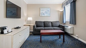 A hotel room with a grey sofa, a wooden coffee table, a desk with a lamp, and a framed picture on the wall. - Glenwood Inn & Suites (Trail)