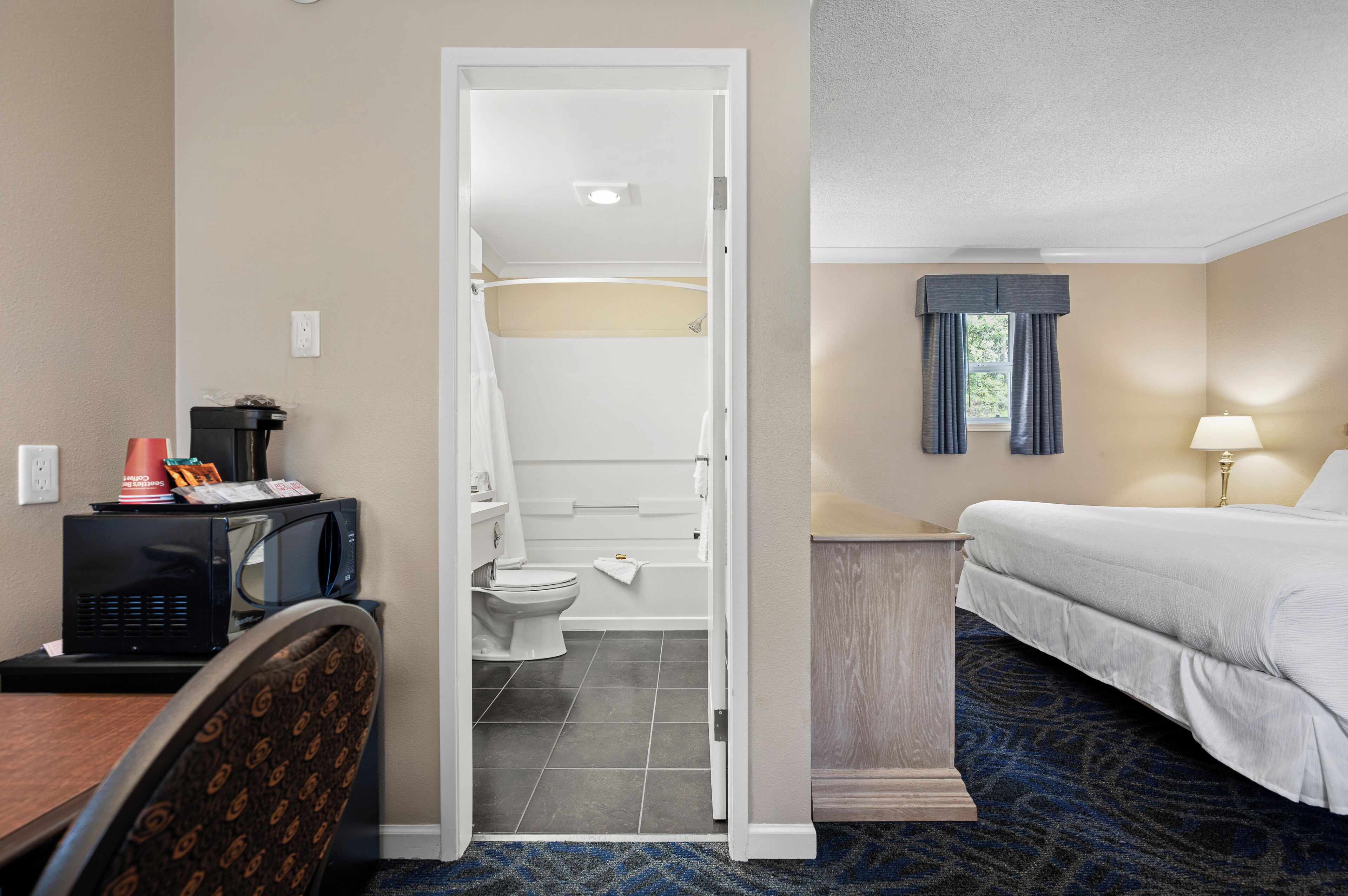A hotel room with a bed, a desk with a microwave, a chair, a bathroom with a toilet and sink, and a window with curtains.