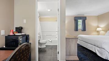 A hotel room with a bed, a desk with a microwave, a chair, a bathroom with a toilet and sink, and a window with curtains.