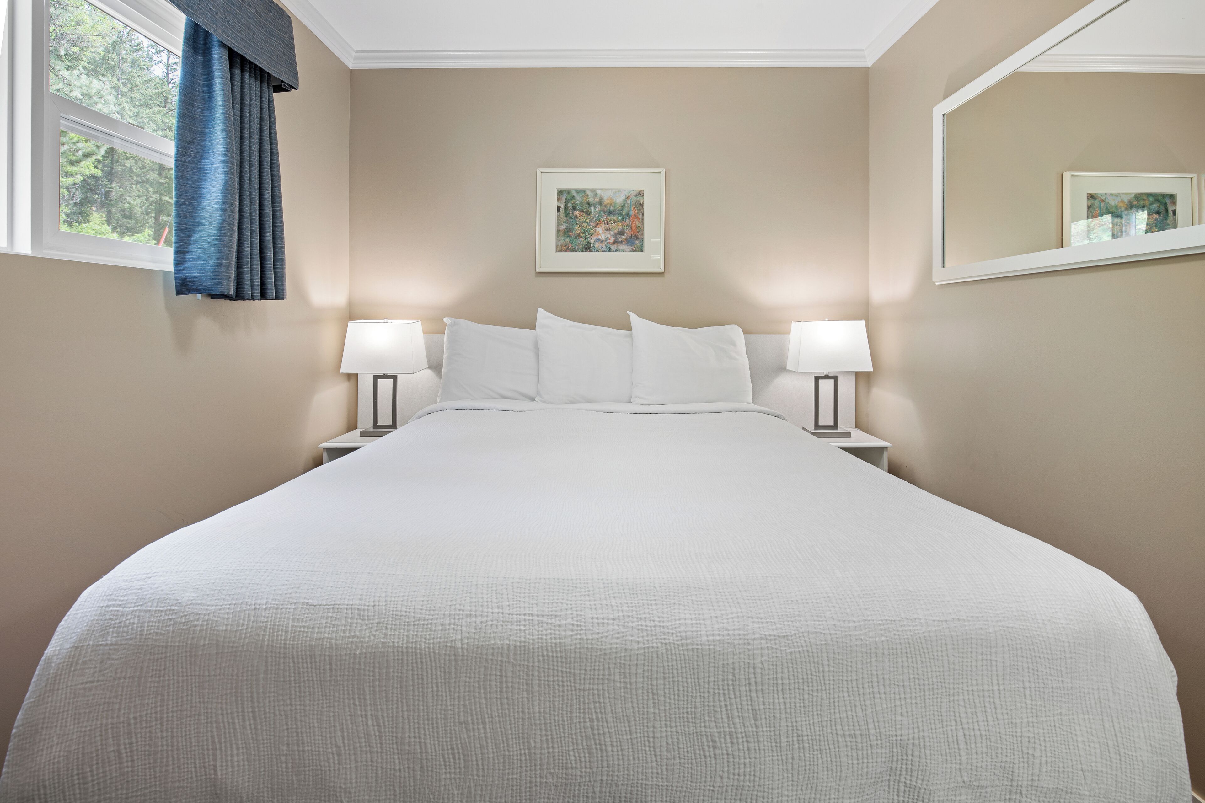 A neatly made bed with white linens, two bedside tables with lamps, a framed picture on the wall, and a window with blue curtains.