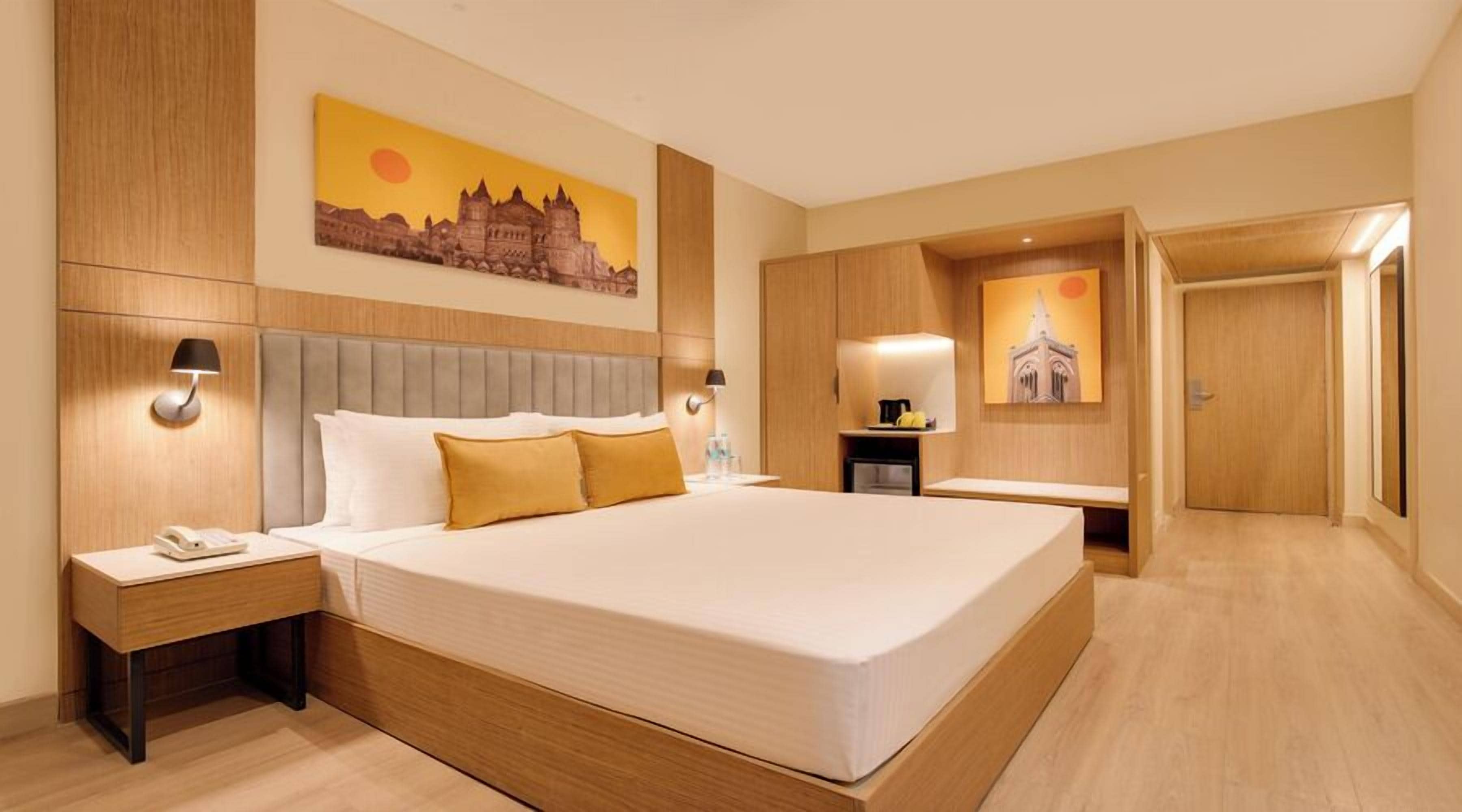 A hotel room with a large bed, bedside tables, a nightstand, a desk, and a chair. There is a painting on the wall and a door leading to another room.