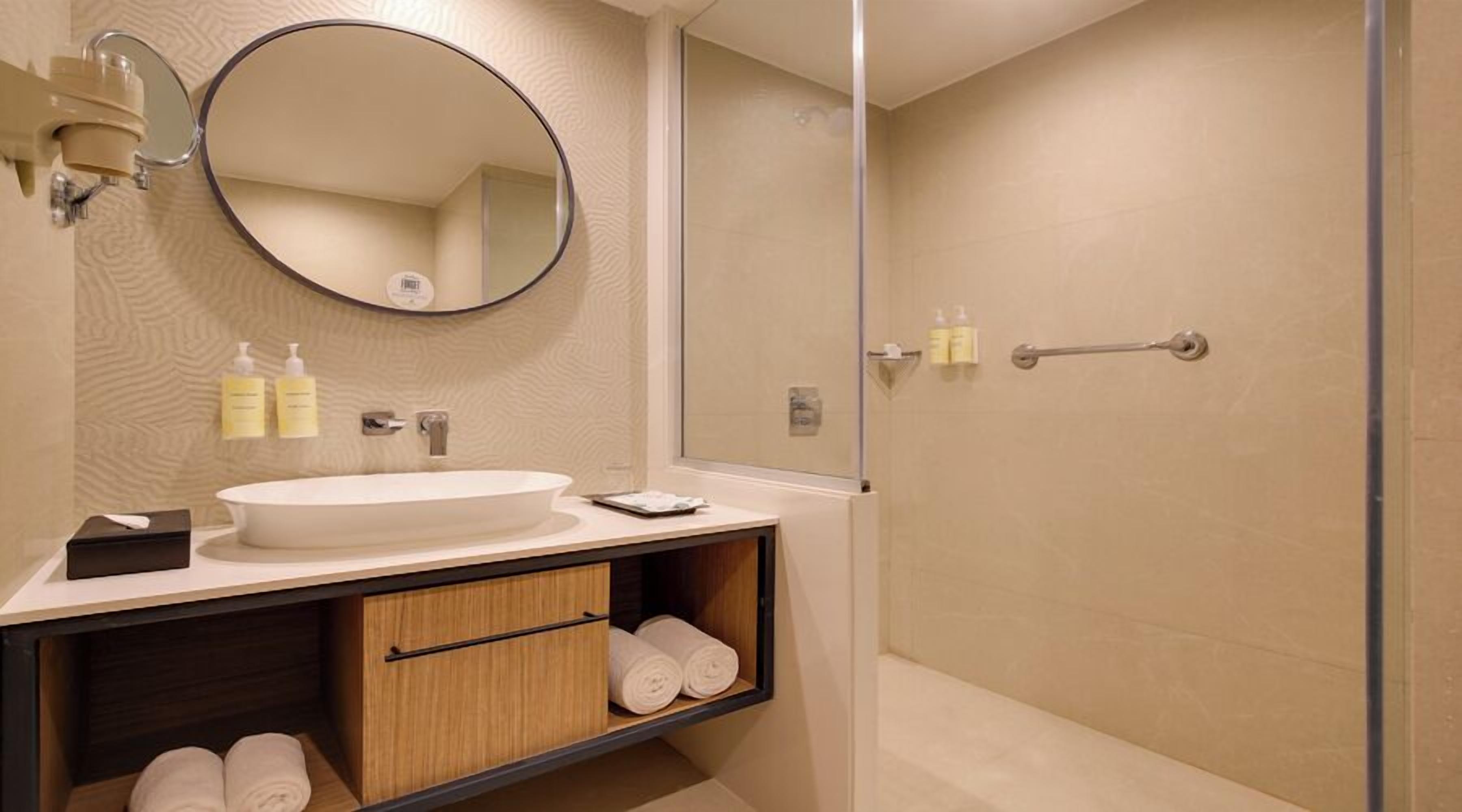 A modern bathroom with a round mirror, a white sink, and a wooden vanity with towels.
