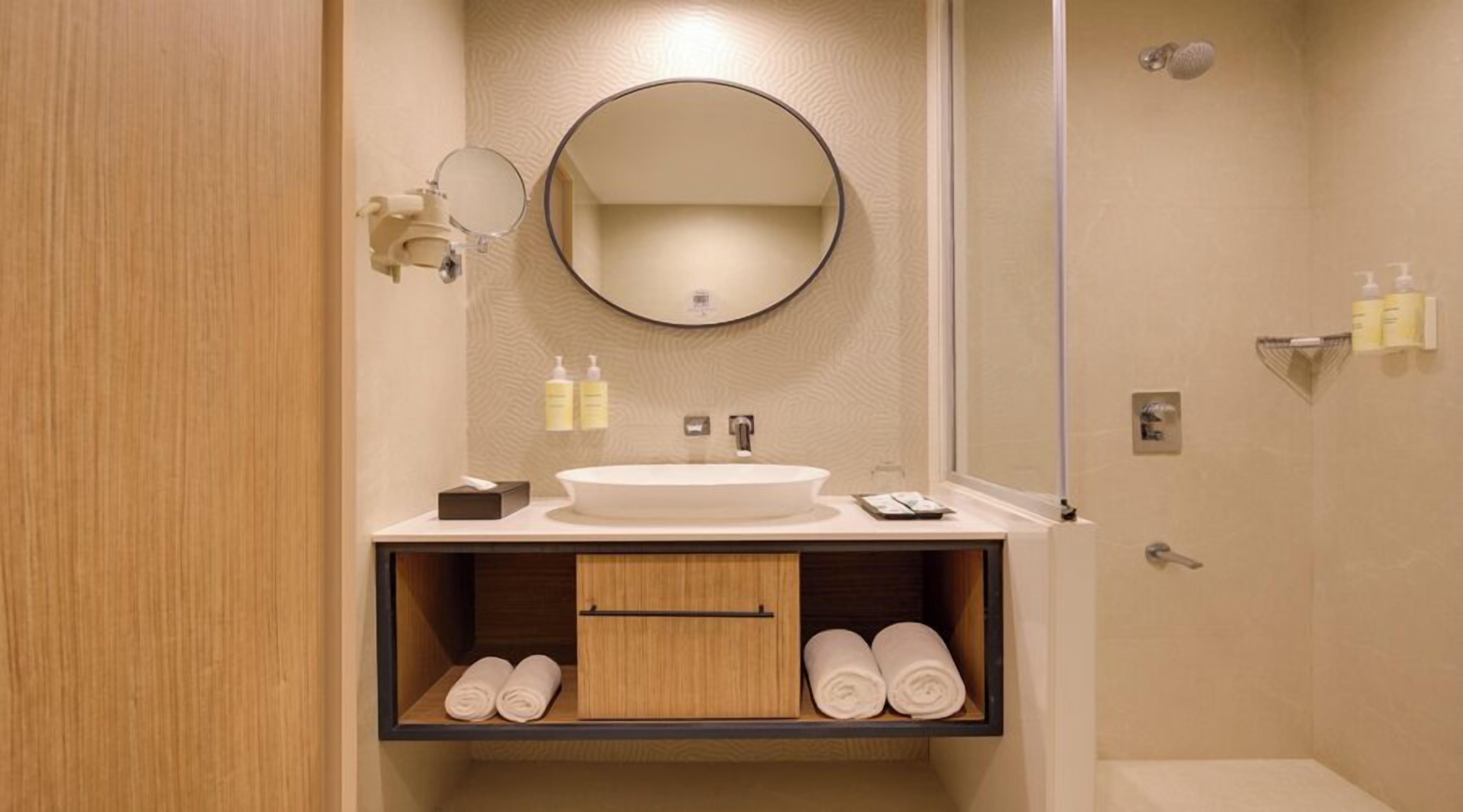 Un baño moderno con un espejo redondo, un lavabo y una ducha. Hay toallas y artículos de aseo en la encimera.
