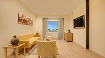 A modern living room with a sofa, armchair, and a table with a vase. A TV is mounted on the wall, and there's a window with curtains.