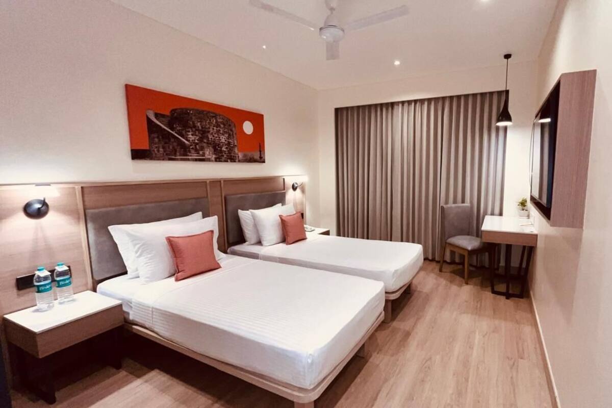 Habitación de hotel con dos camas, un escritorio, una silla, un ventilador de techo y un ventanal grande con cortinas.
