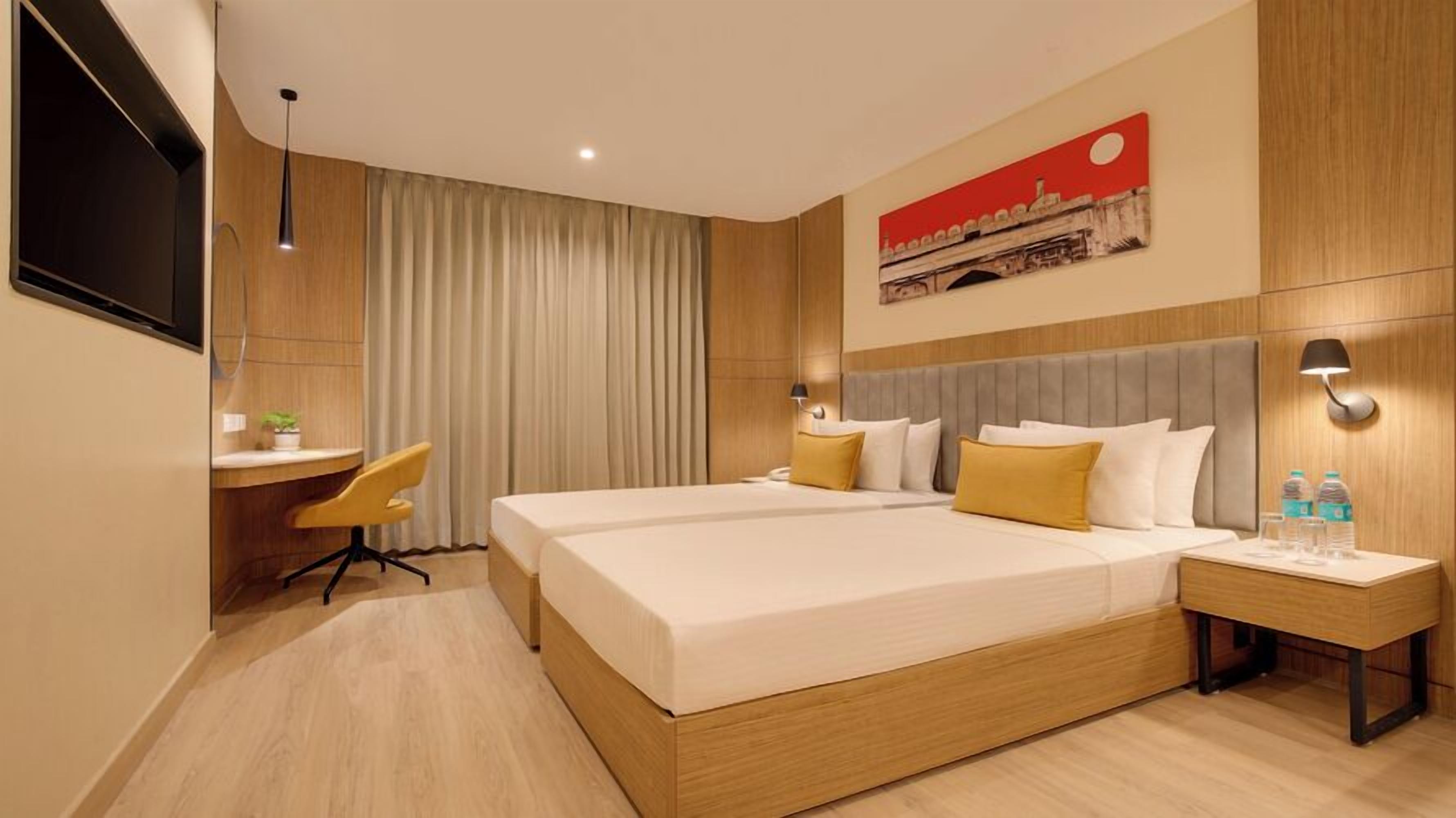 A modern hotel room with a large bed, a desk, and a chair. There is a painting on the wall and a lamp on the nightstand.