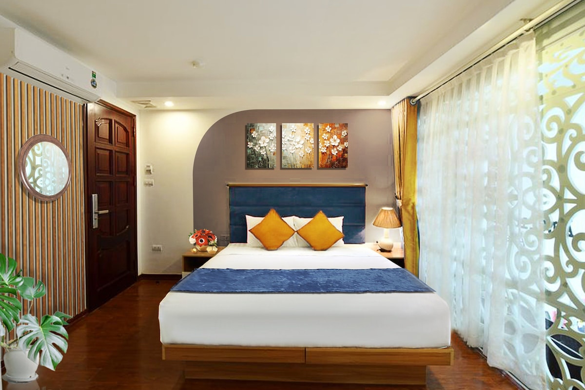 Hotelkamer met een groot bed, houten vloer, een decoratieve spiegel, een deur en een raam met gordijnen.