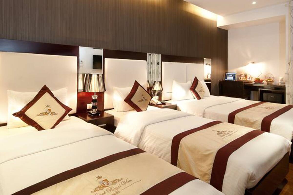 A hotel room with three beds, each with a red and white bedspread and matching pillows. There is a desk with a computer and a lamp, and a window with curtains.