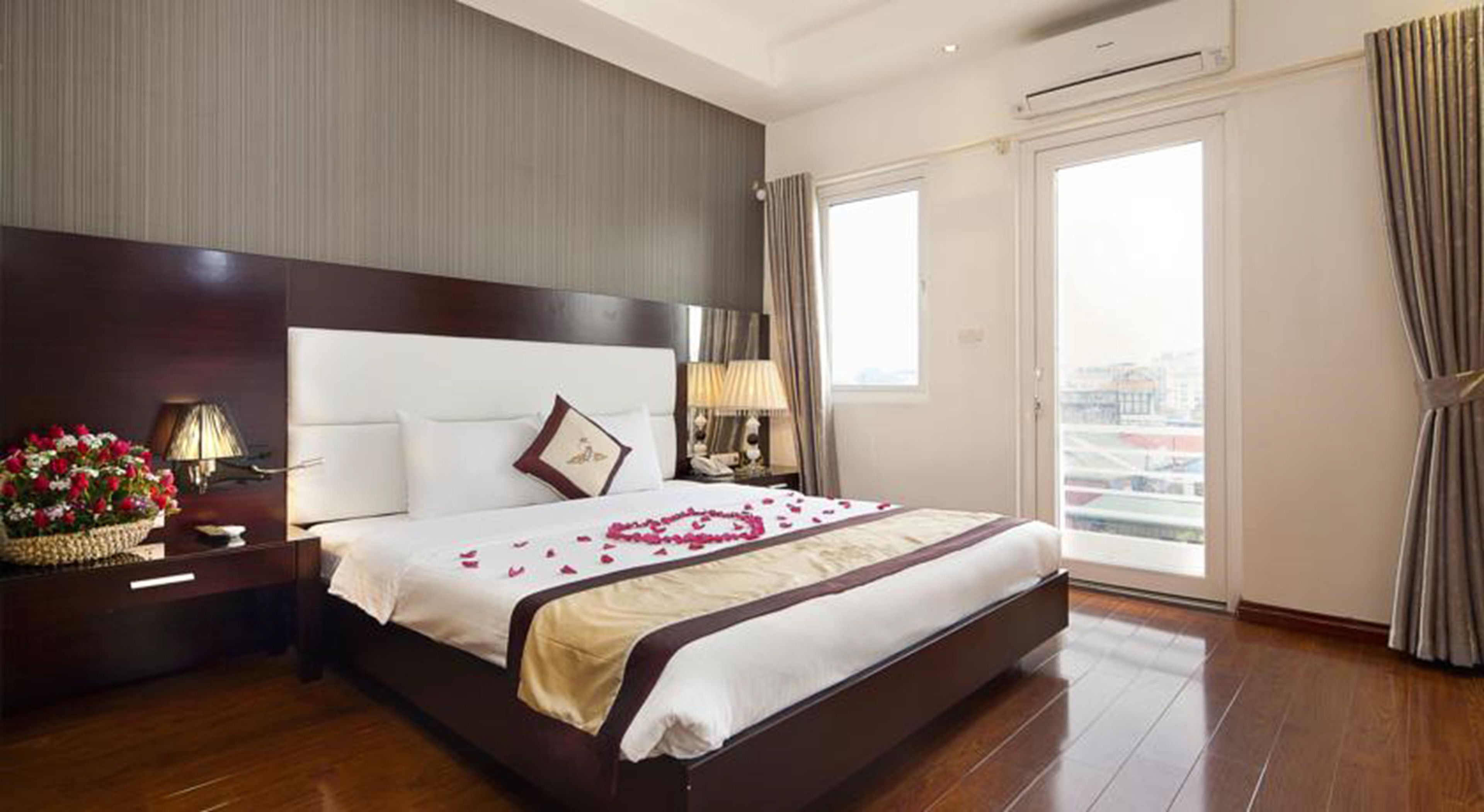 Una habitación de hotel con una cama grande, una mesita de noche con una lámpara, un arreglo floral y vistas a la ciudad a través de la ventana.