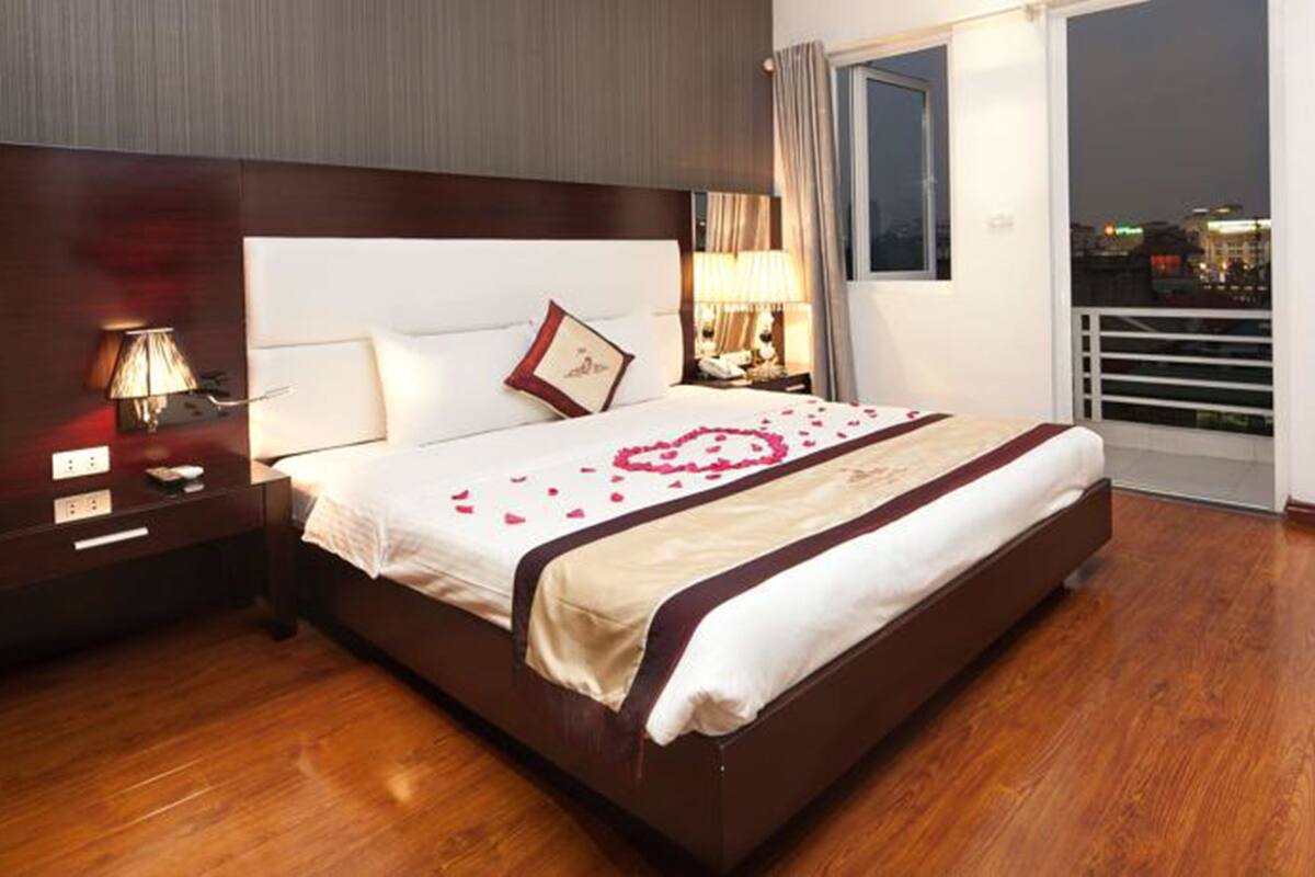 A hotel room with a large bed, a nightstand, a lamp, and a view of the city at night.