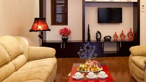 Een woonkamer met twee beige banken, een rode tafel met een fruitschaal, een televisie aan de muur en decoratieve vazen.