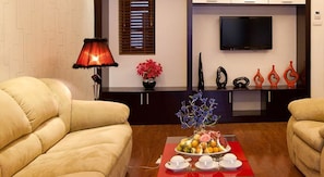 Ein Wohnzimmer mit zwei beigen Sofas, einem roten Tisch mit Obstplatte, einem Fernseher an der Wand und dekorativen Vasen.
