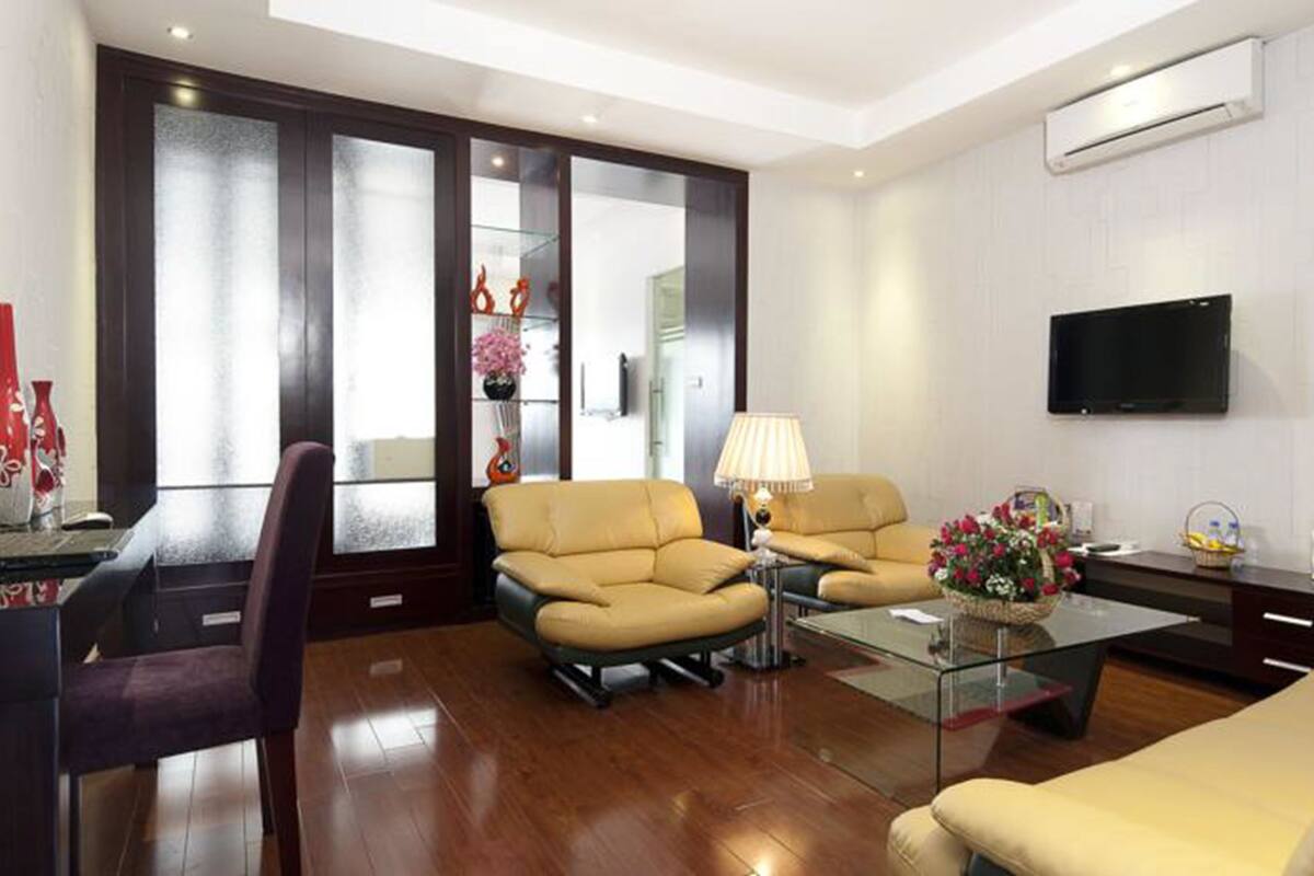 A modern living room with a glass coffee table, two beige sofas, a desk with a laptop, and a TV mounted on the wall.