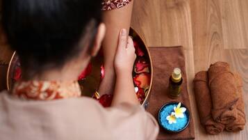 A person receiving a foot soak with rose petals, a bottle of oil, and a blue bowl with flowers on a wooden floor.