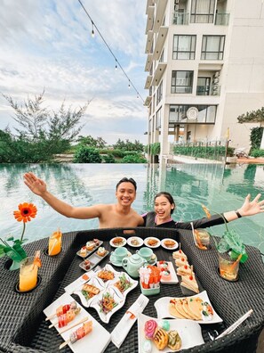 Una mesa de comedor con diferentes platos y bebidas al lado de una piscina, con un edificio al fondo.