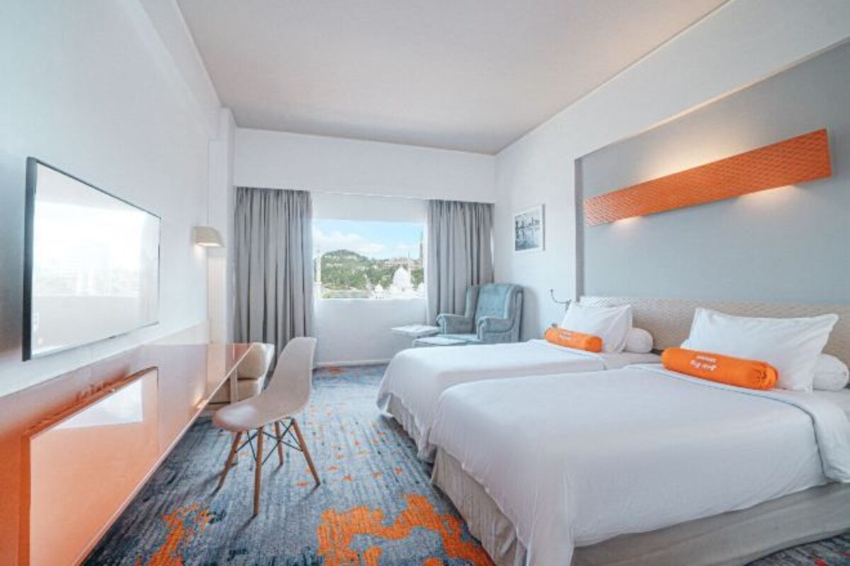 Habitación de hotel con dos camas, un escritorio, una silla y un amplio ventanal con vistas a la ciudad.