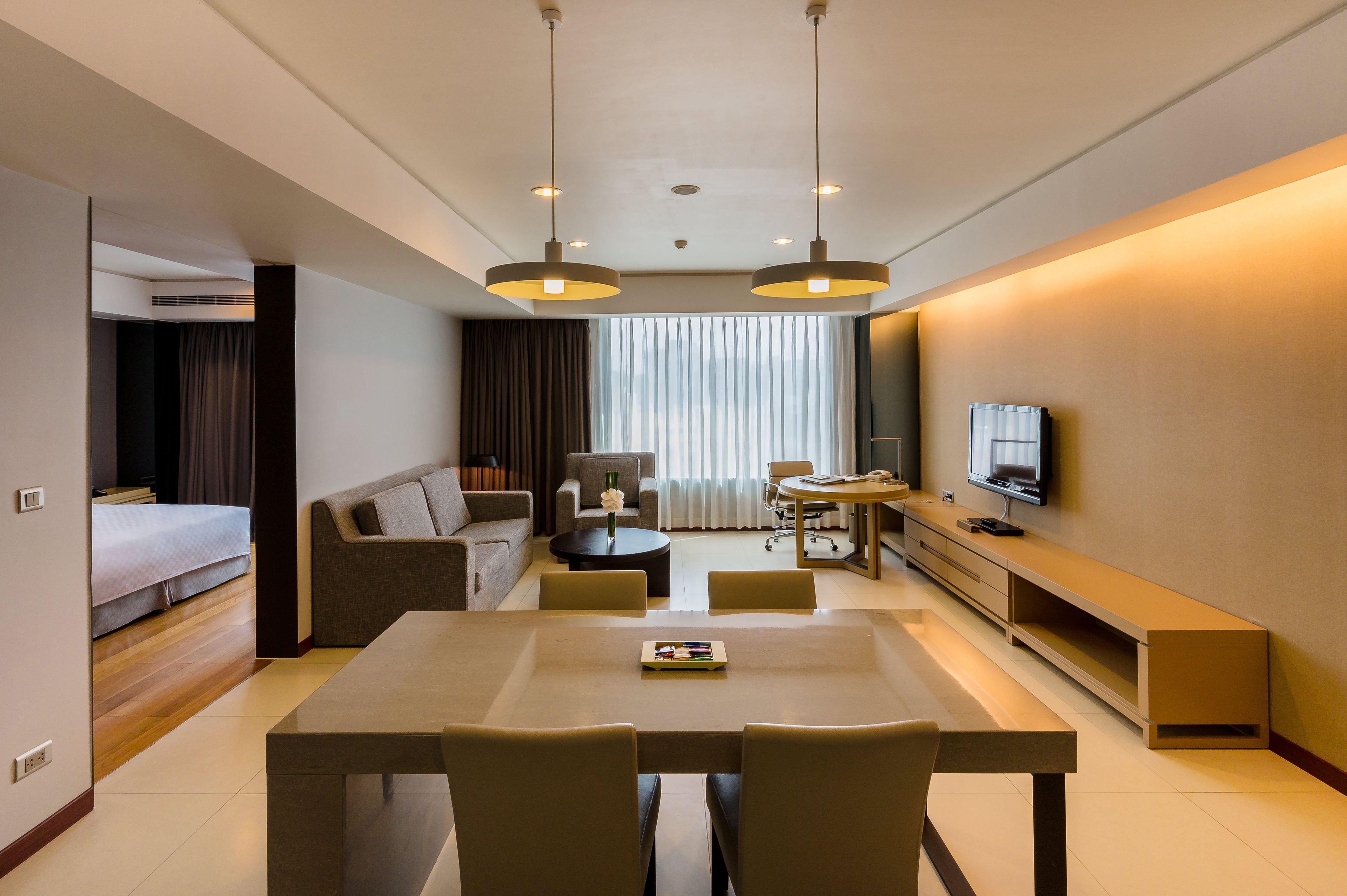A modern hotel room with a dining area, a sofa, a TV, and a large window with curtains.