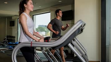 Dos personas corriendo en cintas de correr dentro de un gimnasio con equipo de ejercicio y un ventanal grande al fondo.
