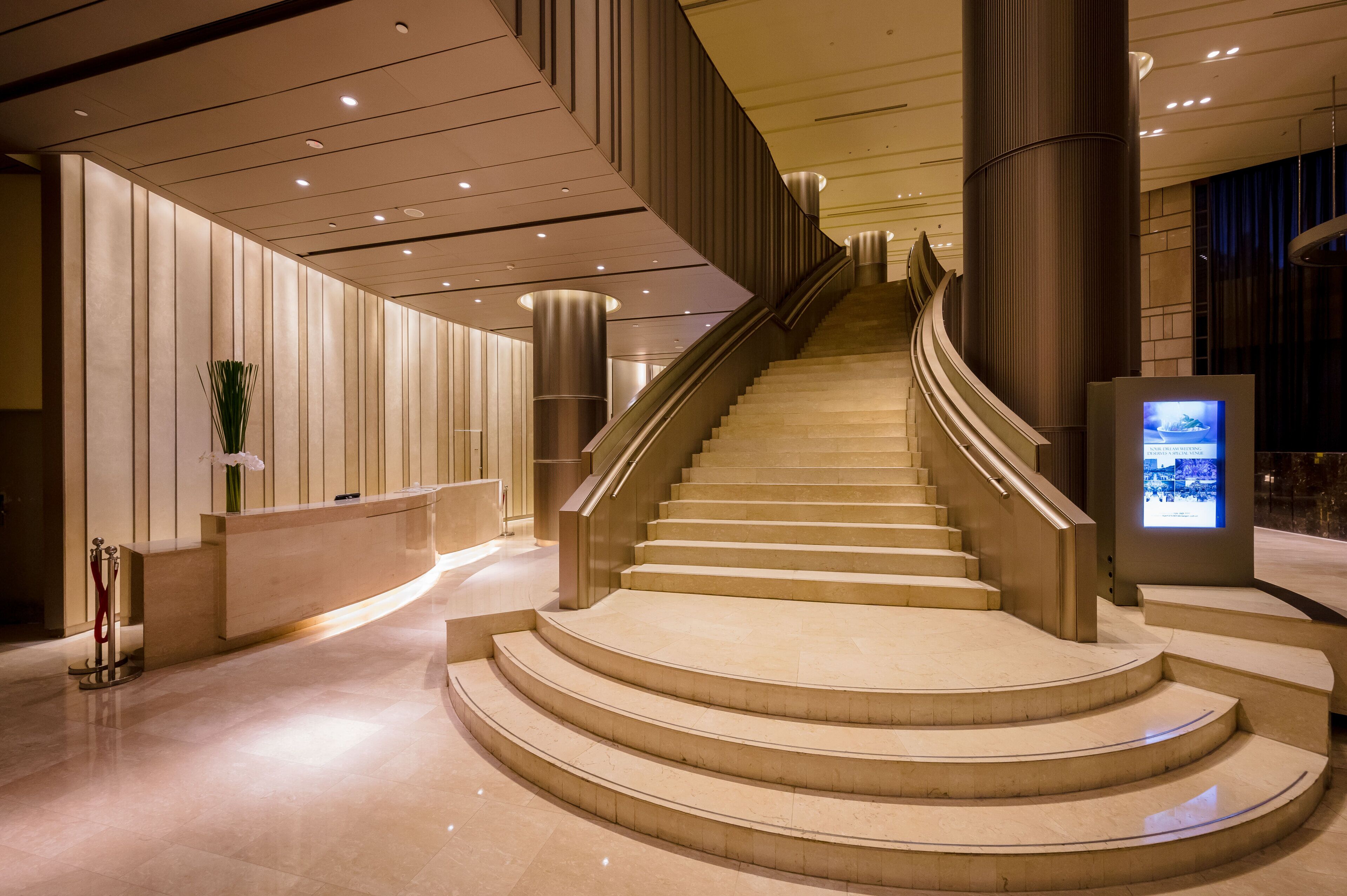 Una gran escalera en un vestíbulo moderno con un mostrador de recepción y una pantalla digital.