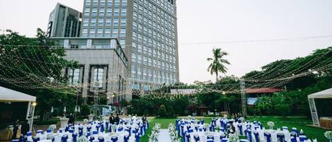 Un lieu de réception en plein air pour un mariage, avec des tables disposées en rangées, un grand bâtiment en arrière-plan et des guirlandes lumineuses suspendues au-dessus.