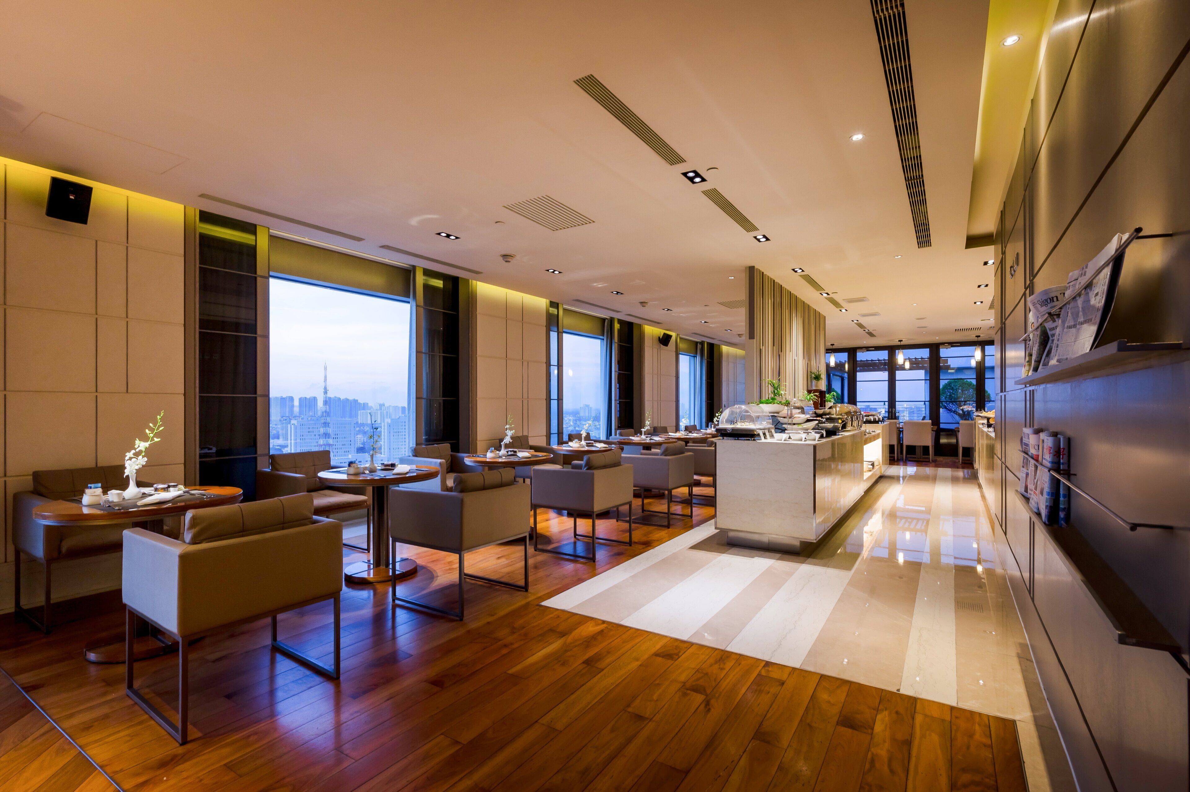 A modern hotel lobby with a wooden floor, a central seating area with sofas and tables, large windows offering a city view, and a kitchen area visible in the background.