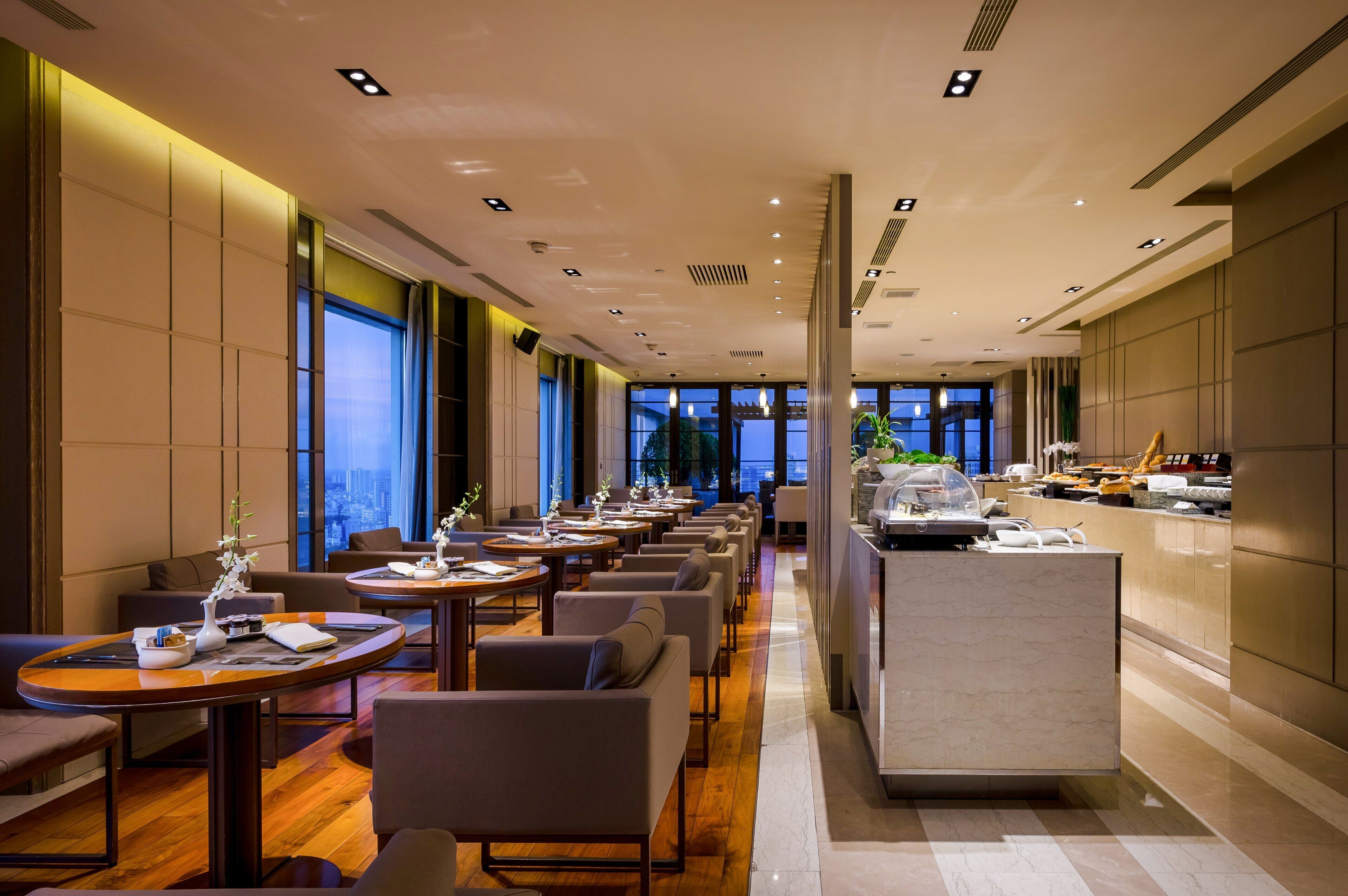 A modern hotel lobby with a long reception desk, multiple seating areas, and large windows offering city views.