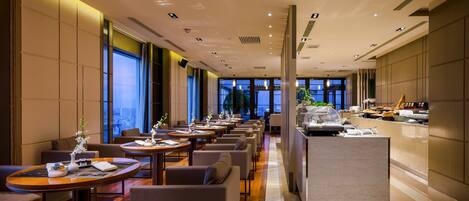 A modern hotel lobby with a long reception desk, multiple seating areas, and large windows offering city views.