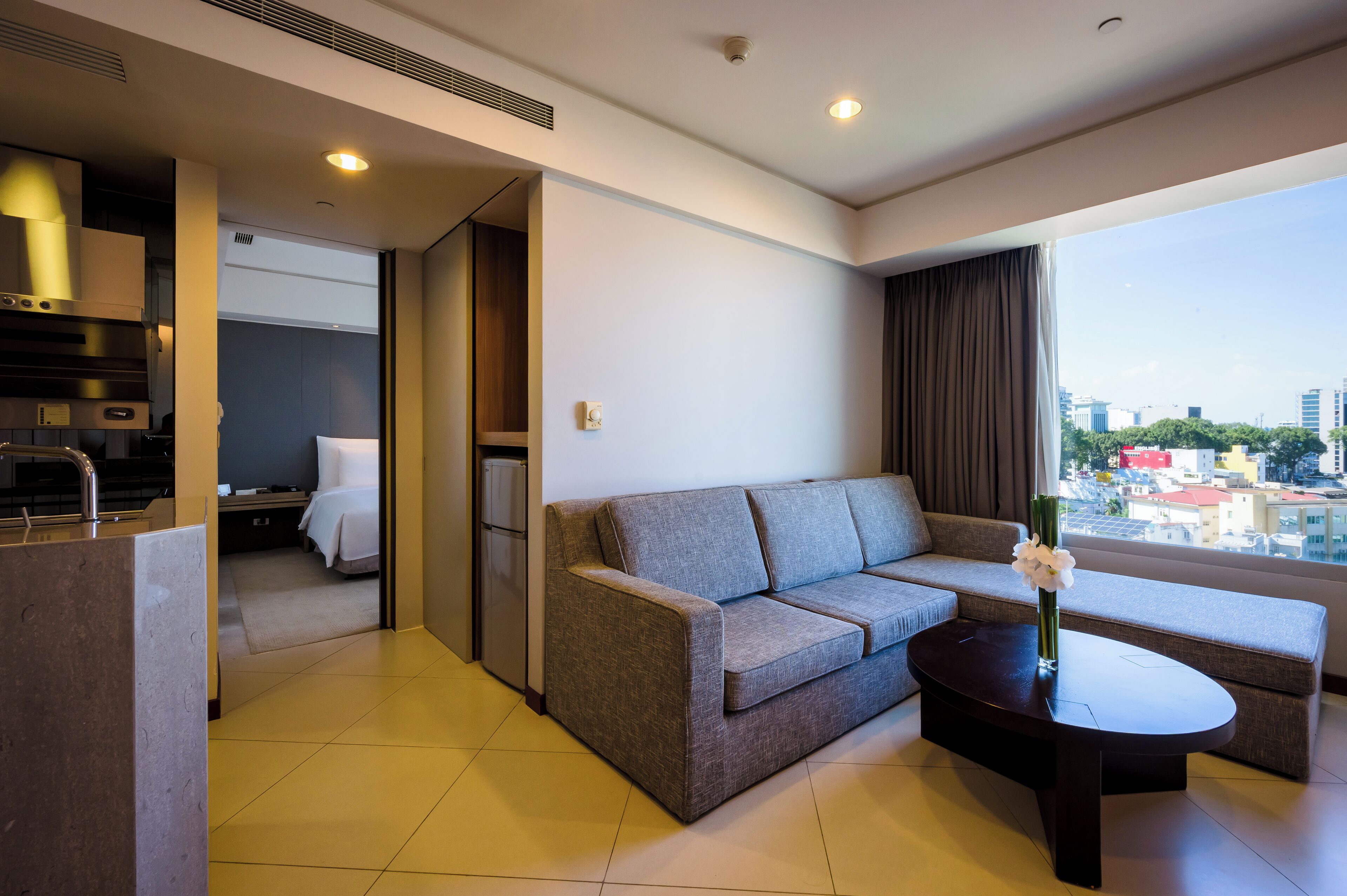 A modern hotel room with a sofa, a coffee table, a view of the city, and a kitchenette.