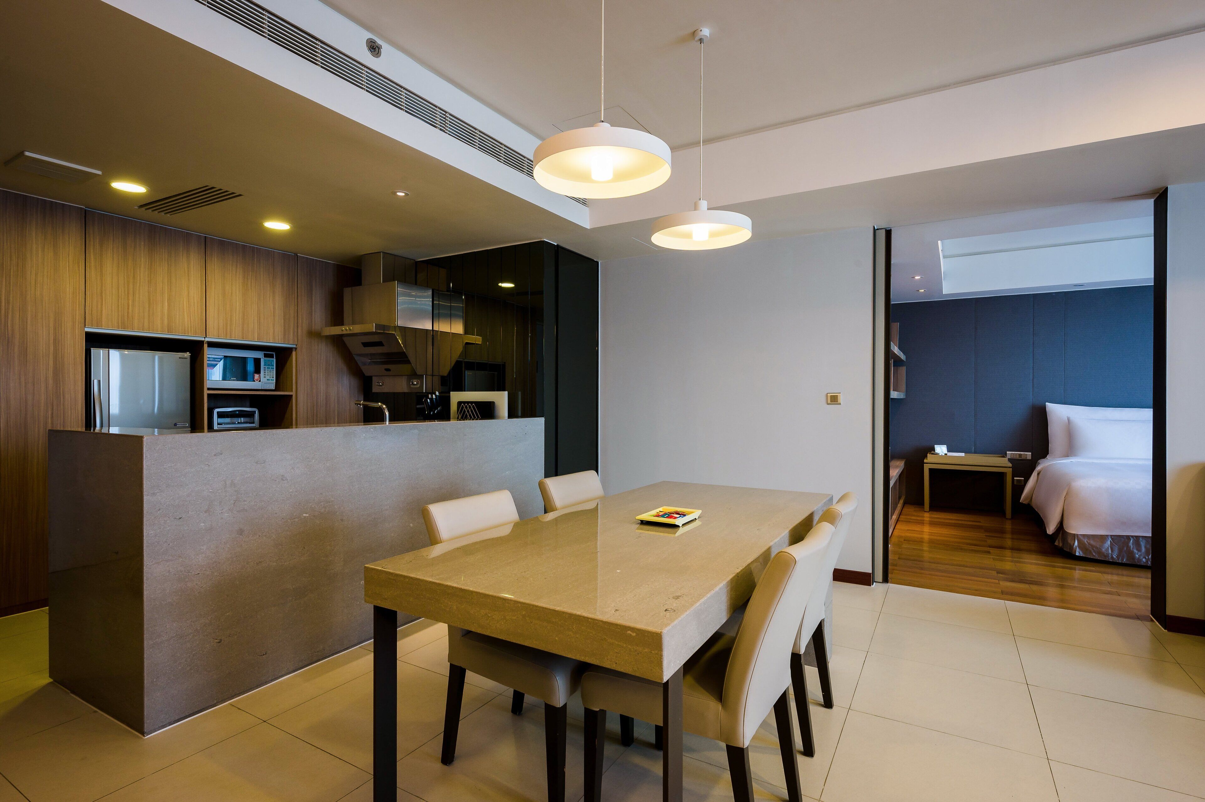 A modern hotel room with a dining area, kitchenette, and a separate bedroom.