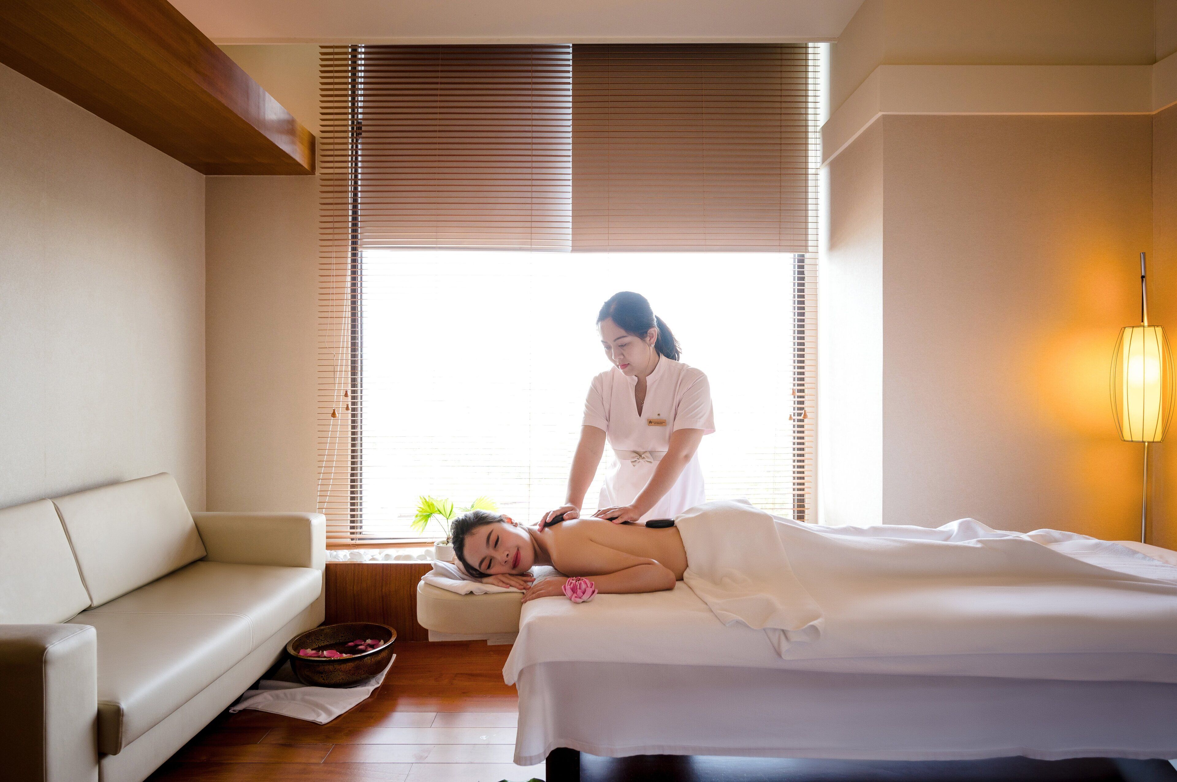 A massage therapy room with a massage table, a sofa, and a window with blinds.