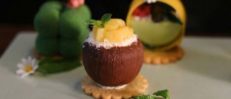 A dessert presentation with a chocolate-covered item topped with fruit and mint, set on a plate with a decorative base.