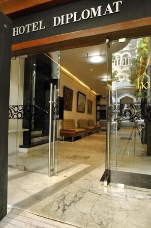 Entrada de um hotel com porta de vidro, área de recepção com assentos e vista para um edifício ao fundo.