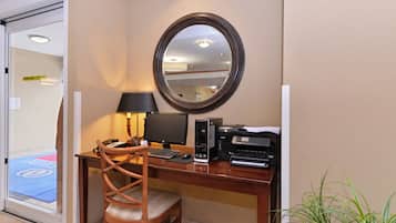 A hotel room with a desk, chair, computer, printer, and a mirror on the wall.