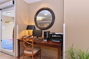 A hotel room with a desk, chair, computer, printer, and a mirror on the wall.