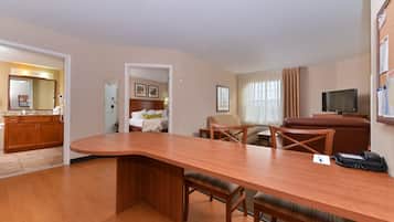 A hotel room with a wooden dining table, a TV, a sofa, and a kitchen area.