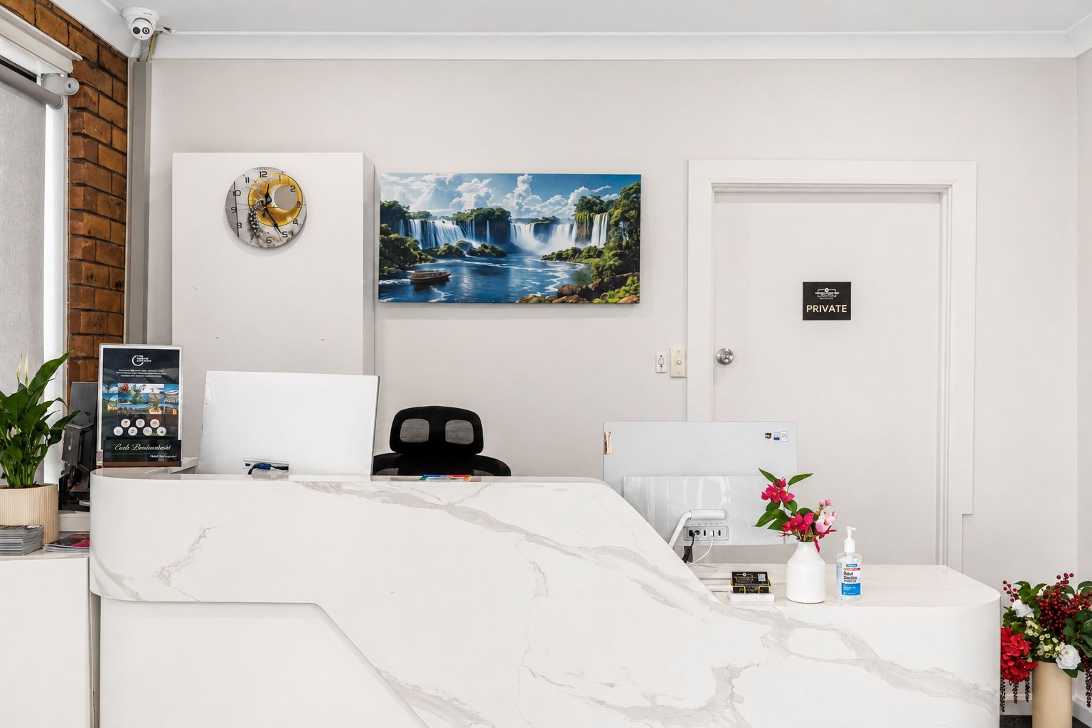A hotel reception area with a marble reception desk, a clock, a painting of a waterfall, a private room sign, and a plant.