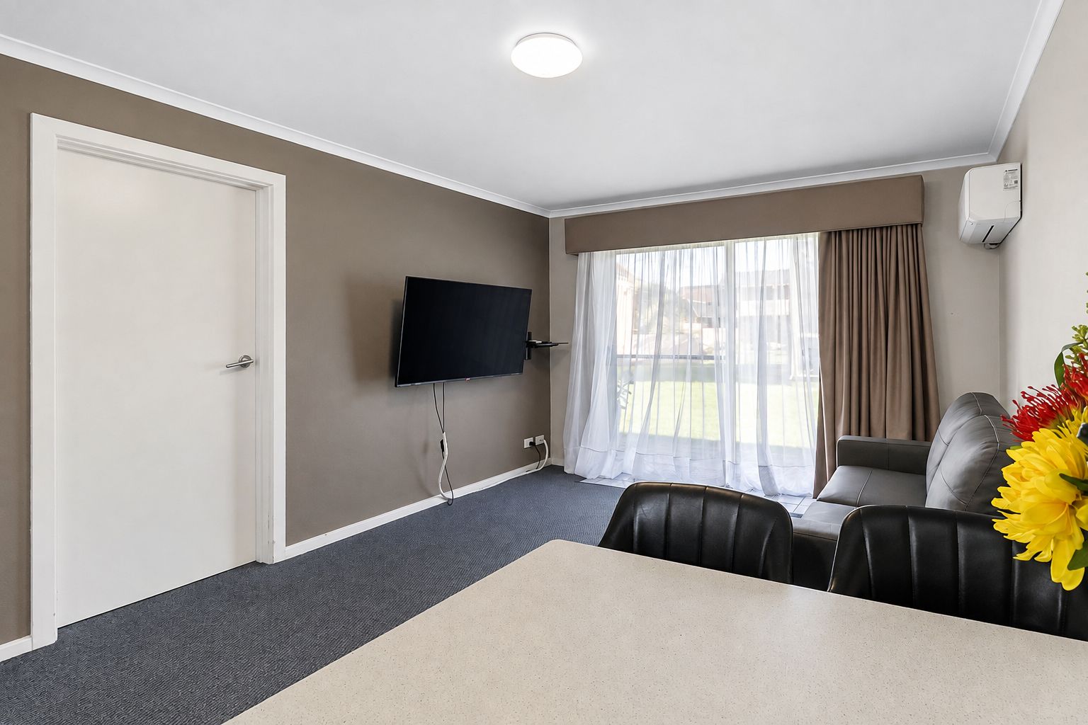 Une chambre d’hôtel équipée d’un téléviseur à écran plat fixé au mur, d’une table à manger avec des chaises, d’un canapé et d’une fenêtre avec des rideaux.