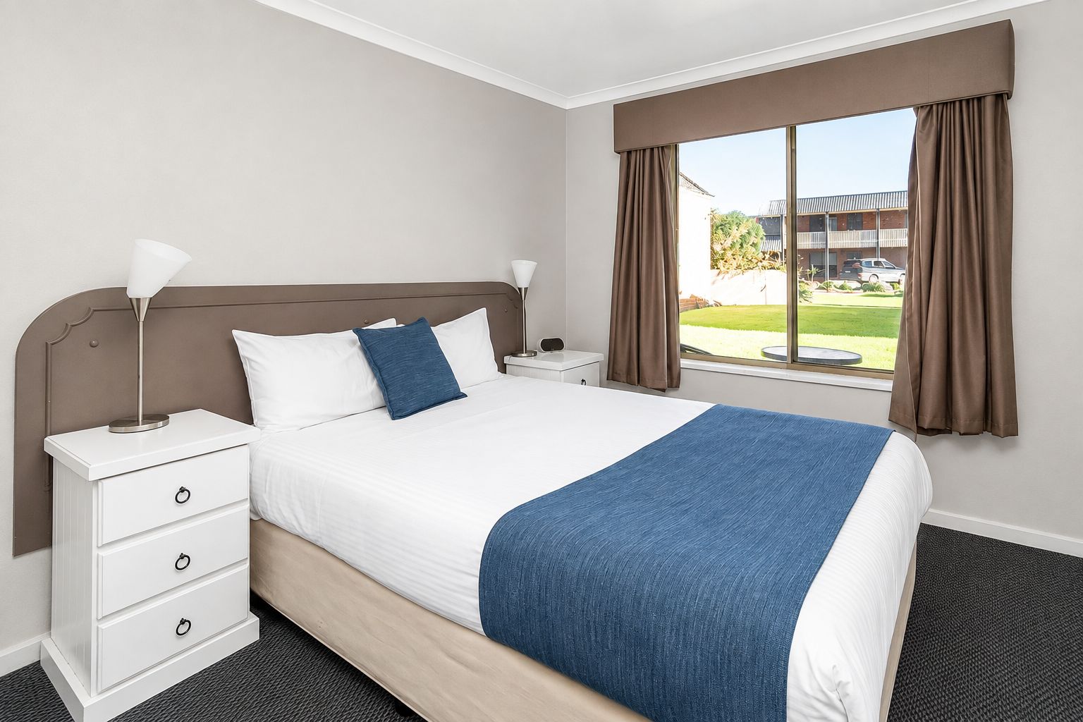 A hotel room with a bed, a nightstand, a lamp, a window with curtains, and a view of the outside.