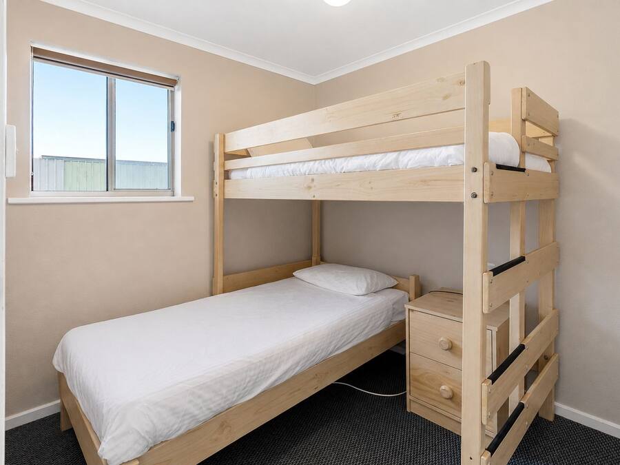 A bunk bed with a single mattress, a bedside drawer, and a window with blinds.