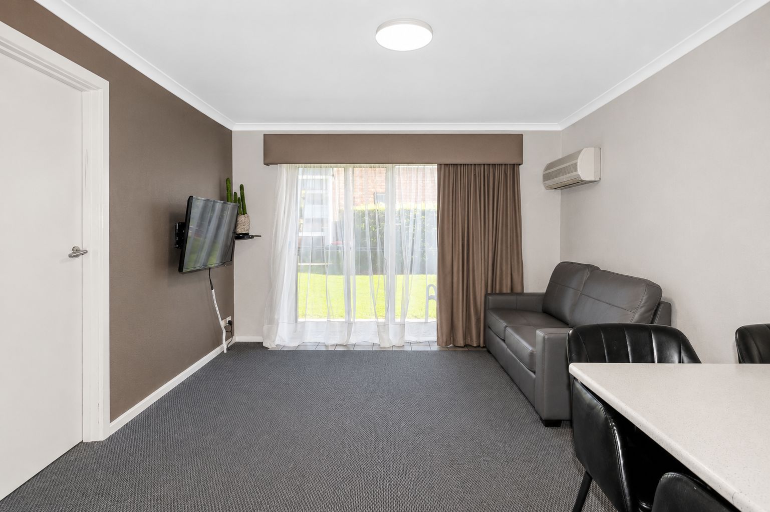 A hotel room with a sofa, a dining table, a TV mounted on the wall, and a large window with curtains.