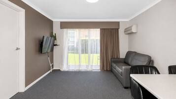 A hotel room with a sofa, a dining table, a TV mounted on the wall, and a large window with curtains.